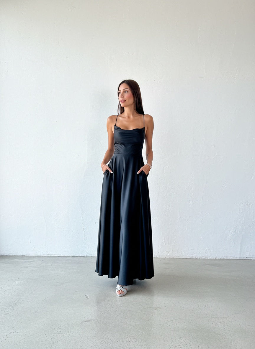 Modelo con vestido largo de satín negro, de tirantes finos, espalda ajustable y falda fluida con bolsillos.